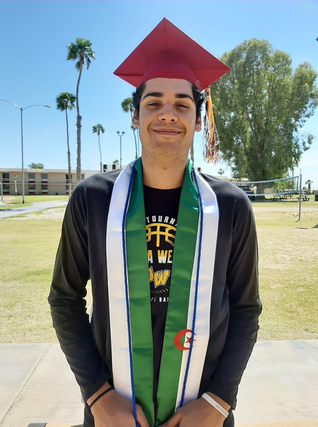 A graduate stands wearing sashes and a cap in front of the AWC dorms