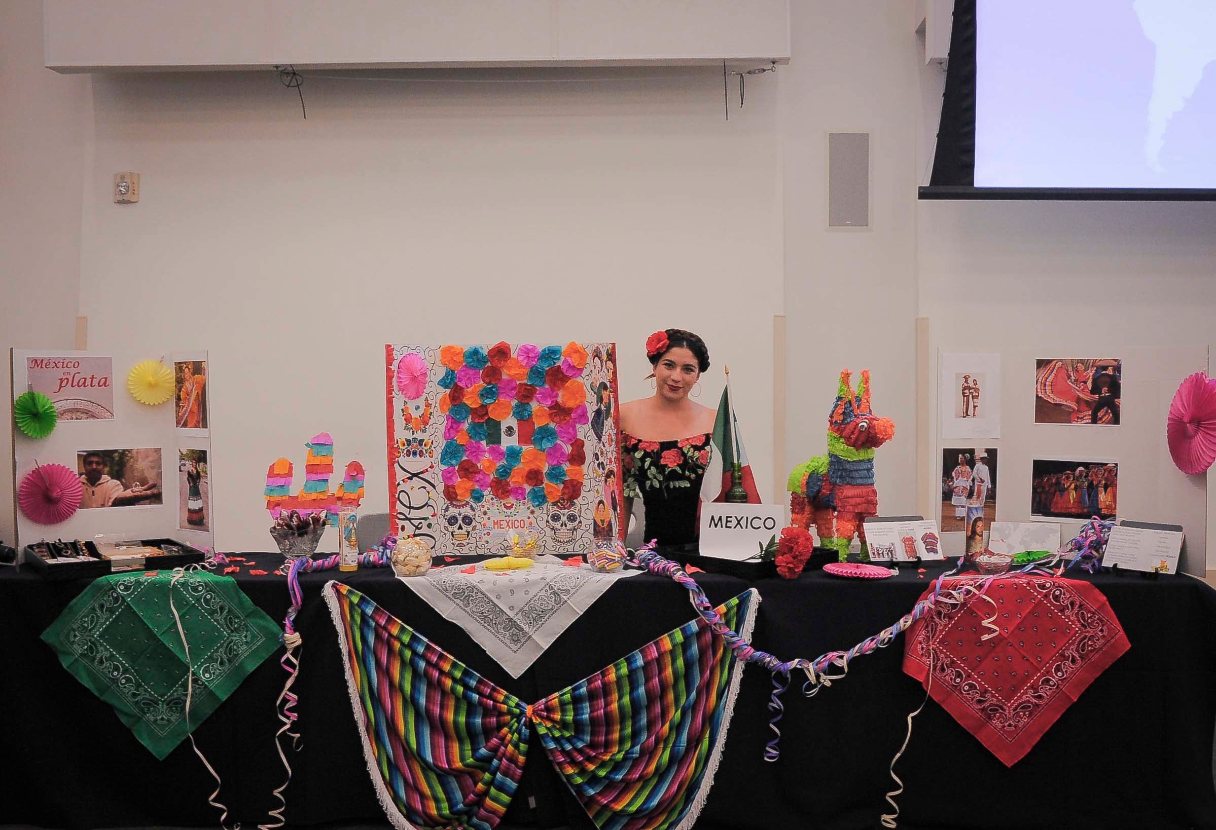 Girl stands behind bright and colorful "Mexico" table. Flowers, a piñata, and paper decor surround her.