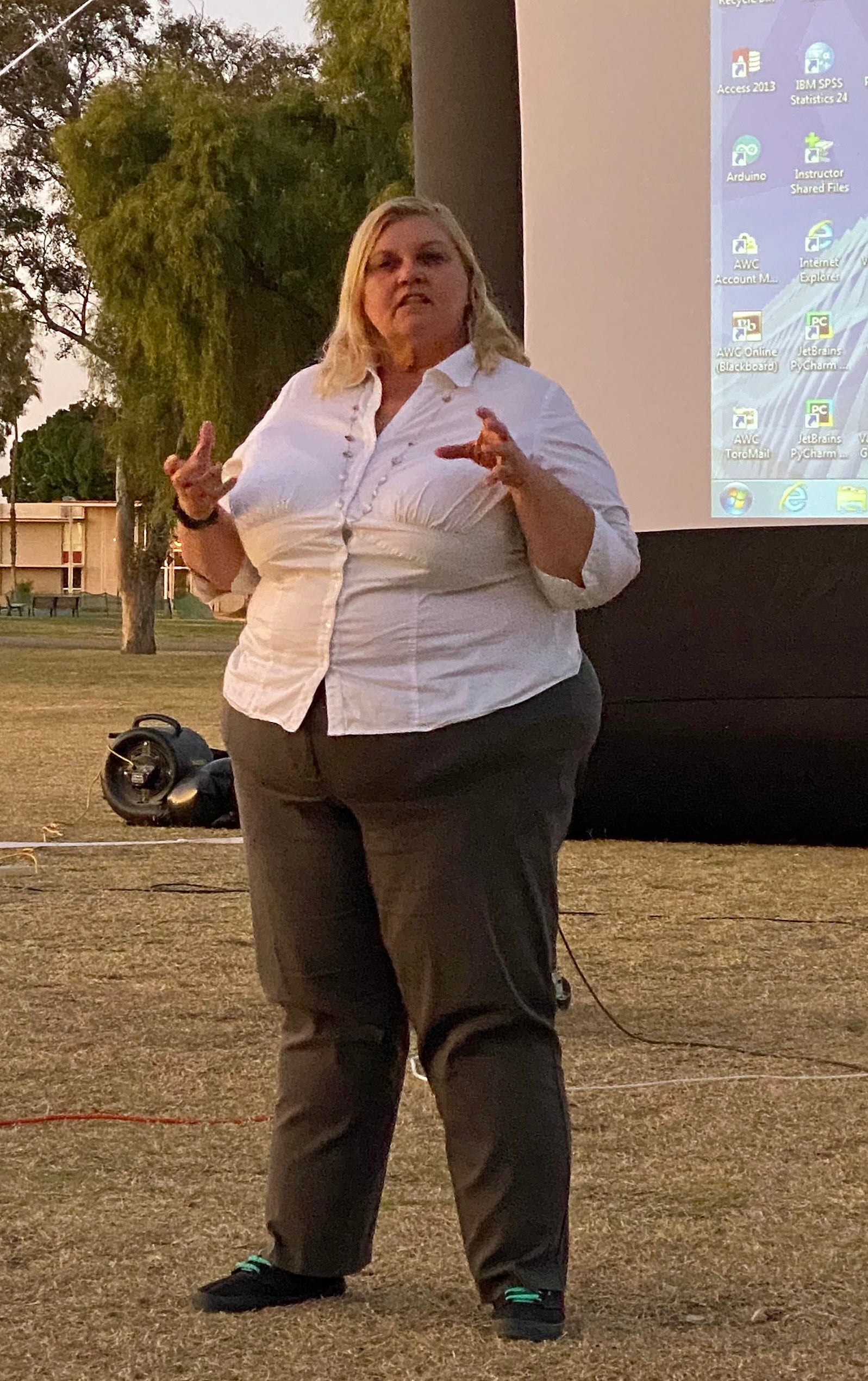 A blonde woman in a white shirt is speaking outdoors in front of a projection screen.