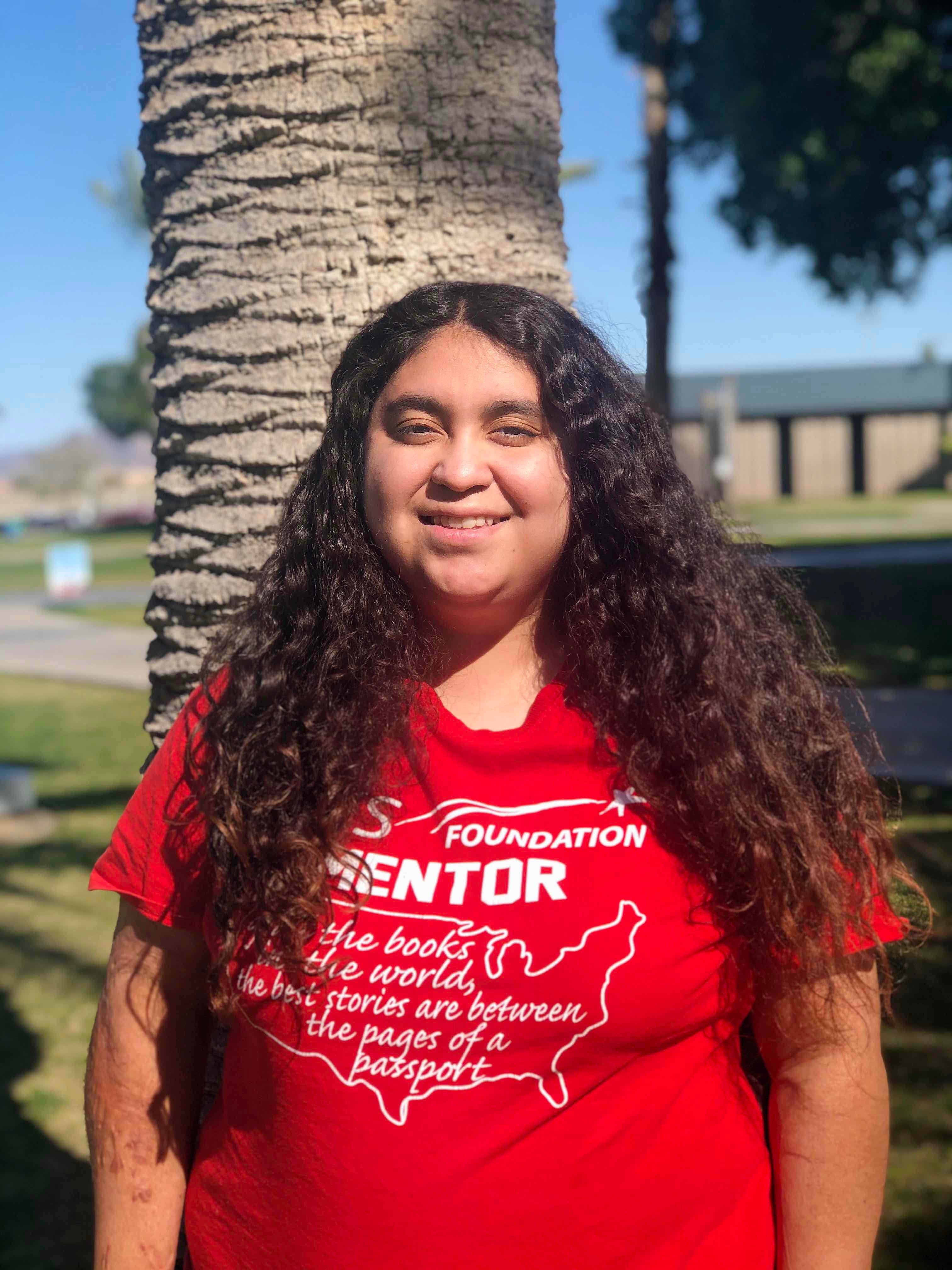 A girl with long black curly hair is wearing a red shirt that says "Foundation Mentor" and is smiling in front of a palm tree.