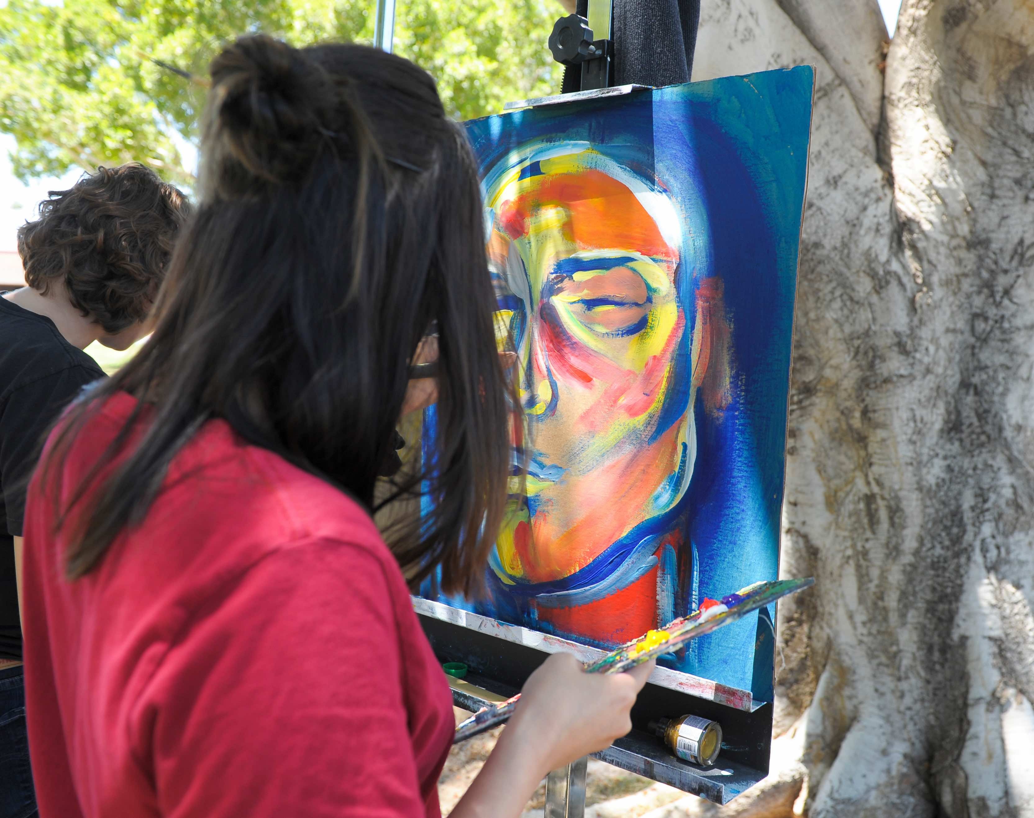 Students paint during the March 2018 Student Art Exhibition Reception on the Yuma Campus. Photo by Alex Lastra, AWC Photo Services. A person in red is standing outside painting on a canvas which is already blue with an orange face