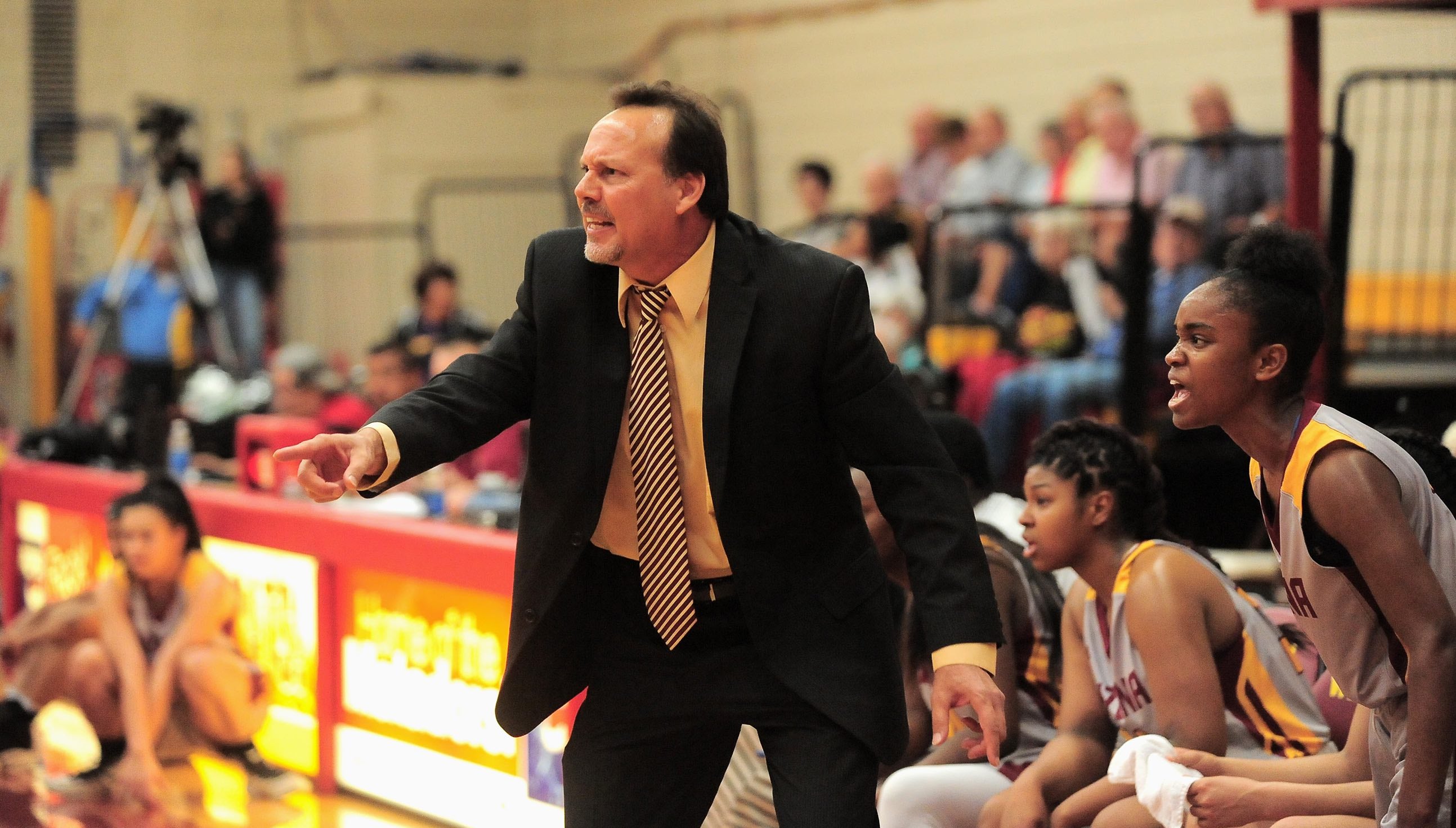 Coach Patrick Cunningham leading his team during a Region 1 playoff game against Eastern Arizona in March 2017. Photo by Alex Lastra, AWC Photo Services. Coach Cunningham is dressed in a black suit and tie, seen yelling towards off-frame players. He is standing inside a sports game in the Basketball court
