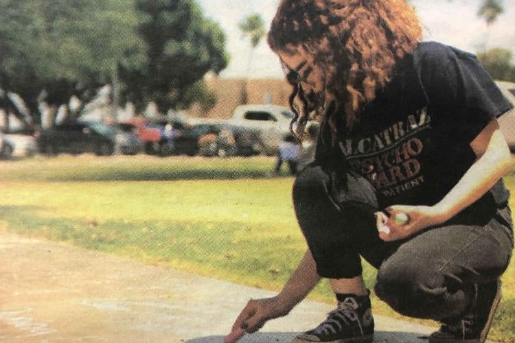 An AWC student writes "gregueria" on campus sidewalk at the Student Art Exhibit Reception in May. Photo Courtesy of AWC Photo Services. Student kneels down to write with chalk on school sidewalk