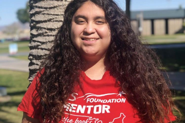 A girl with long black curly hair is wearing a red shirt that says "Foundation Mentor" and is smiling in front of a palm tree.