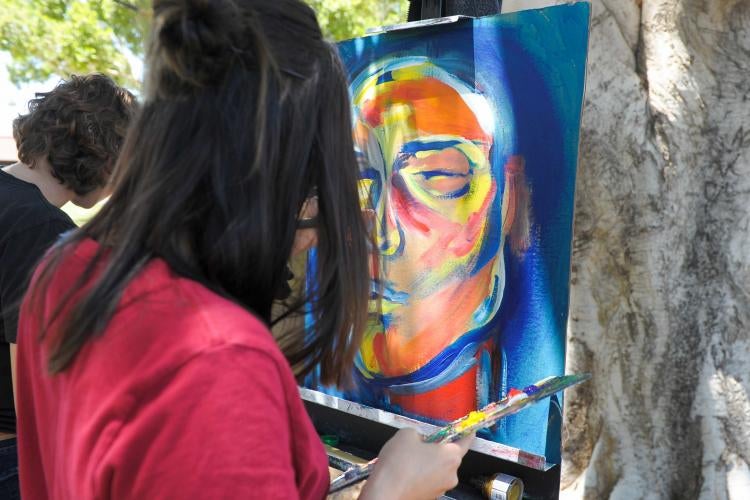 A person in red is standing outside painting on a canvas which is already blue with an orange face 