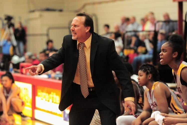 Coach Cunningham is dressed in a black suit and tie, seen yelling towards off-frame players. He is standing inside a sports game in the Basketball court