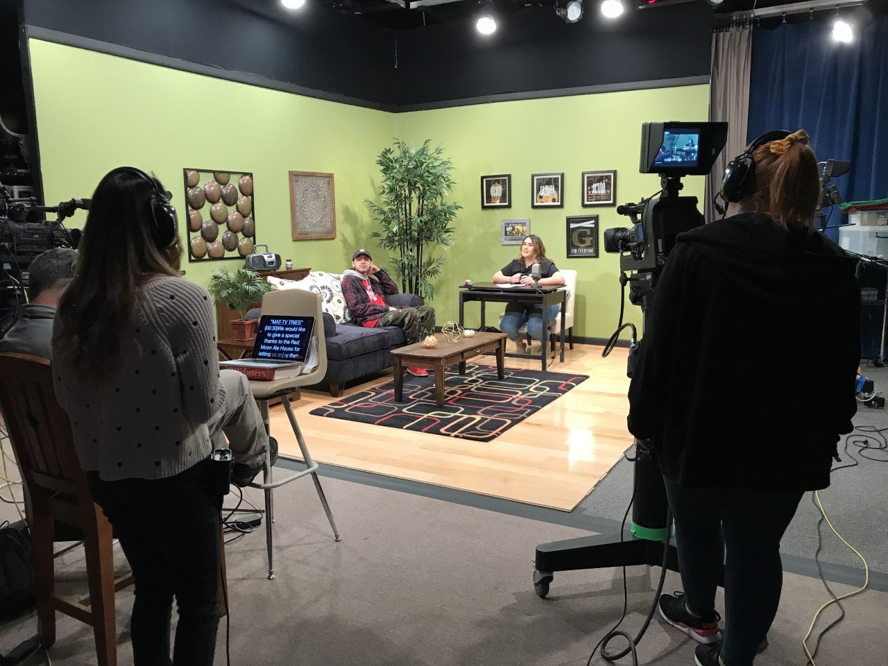 The studio stage--a green room with a coffee table and seats--is foregrounded by camera people.